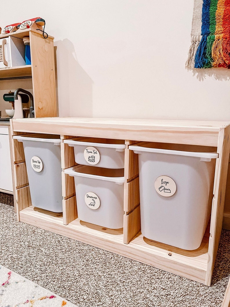 Pu&ograve; includere: Un'unit&agrave; di stoccaggio in legno con quattro contenitori bianchi. I contenitori sono etichettati con cerchi di legno che dicono "Dress Up", "Train Set", "Dinosaurs" e "Lego".