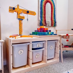 Pu&ograve; includere: Un'unit&agrave; di stoccaggio in legno con tre contenitori etichettati "Macchine", "Musica" e "Travestimento". I contenitori sono bianchi e hanno un'etichetta in legno con testo nero. L'unit&agrave; &egrave; realizzata in legno naturale e ha una finitura in legno chiaro. Una gru giocattolo gialla &egrave; sopra l'unit&agrave;. Un arazzo da parete arcobaleno &egrave; sullo sfondo.