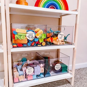 Pu&ograve; includere: Un'unit&agrave; a scaffale bianca con quattro contenitori di plastica trasparente pieni di diversi tipi di giocattoli. I contenitori sono etichettati "Bristle Blocks", "Dinosauri", "Blocchi" e "Magnetic Tiles". Un arco di legno arcobaleno &egrave; sullo scaffale superiore.