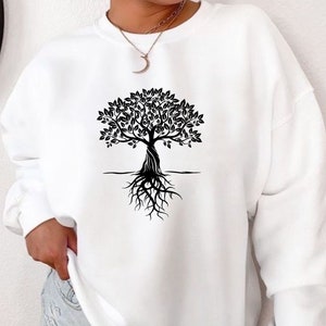 May include: White sweatshirt with a black tree design. The tree has a detailed trunk, leafy branches, and exposed roots. The design is centered on the front. The sweatshirt has long sleeves and a crew neck.