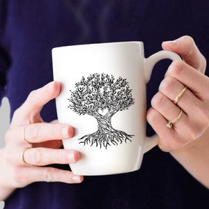 May include: White ceramic mug with a black and white tree design, including a heart-shaped cutout. The tree has detailed roots and branches. The mug is held by a person wearing rings.