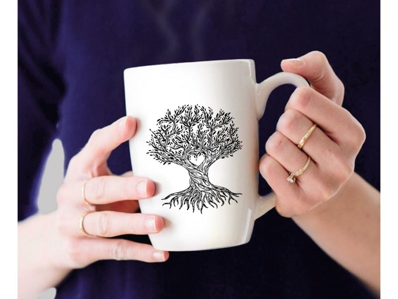 May include: White ceramic mug with a black and white tree design, including a heart-shaped cutout. The tree has detailed roots and branches. The mug is held by a person wearing rings.