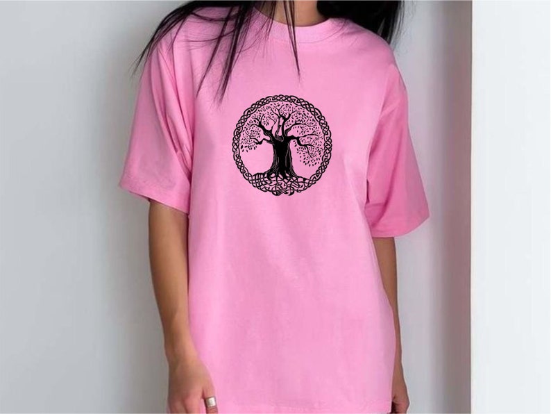 May include: A pale pink t-shirt with a black tree of life design inside a circular Celtic knot border. The short-sleeved shirt is made of a soft material.
