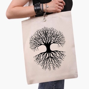 May include: A beige canvas tote bag with a black tree of life design. The tree graphic is symmetrical, with branches and roots forming a circular shape. The bag has two handles and is held by a person.