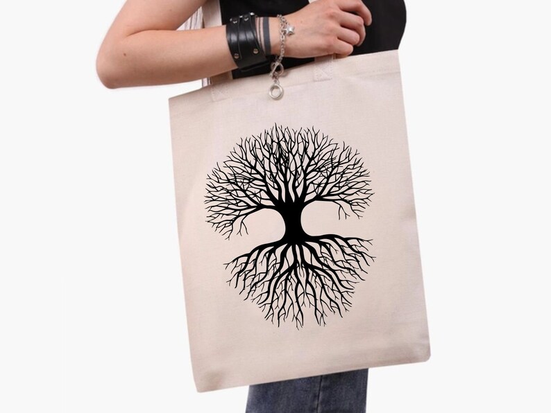 May include: A beige canvas tote bag with a black tree of life design. The tree graphic is symmetrical, with branches and roots forming a circular shape. The bag has two handles and is held by a person.