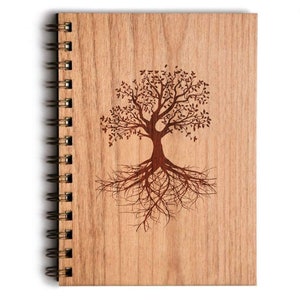 May include: A wooden notebook with a spiral binding, featuring a laser-etched tree design. The tree has a detailed trunk, branches, leaves, and roots. The cover is made of light-coloured wood, and the spiral binding is a dark metal.