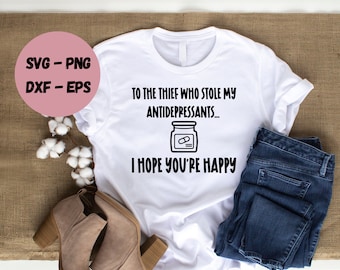 Puede incluir: Camiseta blanca con texto negro que dice "Al ladrón que robó mis antidepresivos... Espero que seas feliz". Una ilustración en blanco y negro de un frasco de pastillas está por encima del texto.