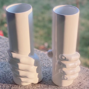 May include: Two white ceramic vases with a unique design. Each vase has a cylindrical shape with a hand-like sculpted detail around the base. The vases are displayed outdoors, with natural light enhancing their smooth, glossy finish.