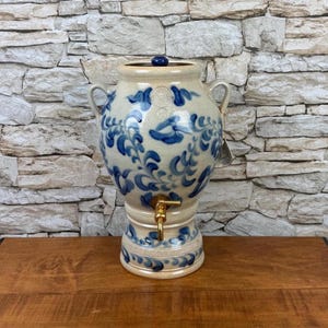 May include: A beige ceramic beverage dispenser featuring a hand-painted blue floral pattern, two side handles, a dark blue lid knob, and a brass-colored metal spigot. The vessel has a wide body and a pedestal base with decorative blue scrollwork.