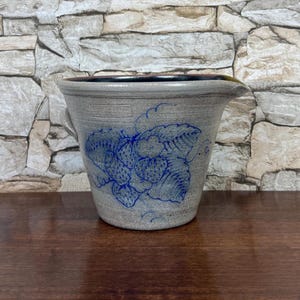May include: A gray stoneware crock featuring a blue hand-painted strawberry motif on the front. The vessel has a wide, flared rim, a small pouring spout on the right side, and a side handle. The interior is dark, and the exterior has a textured finish.