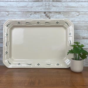 May include: A rectangular, off-white ceramic serving platter with a decorative border. The border features a repeating pattern of small green floral and curved line designs. A small potted plant sits to the right of the platter.