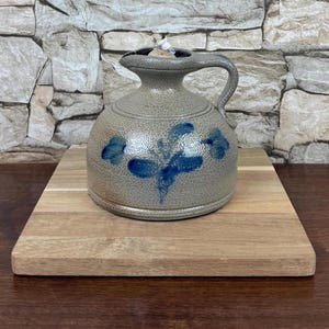 May include: A stoneware pitcher with a handle and a spout, featuring blue floral designs. The pitcher is gray with a speckled texture and sits on a wooden cutting board. The background is a stone wall.