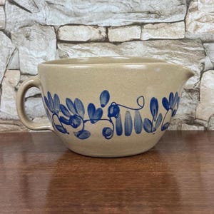 May include: Tan ceramic mixing bowl with a handle and spout, decorated with blue floral patterns. The bowl is set on a wooden surface against a stone wall background. A classic kitchenware item.