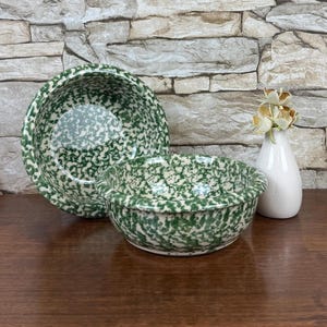 May include: Two green and white speckled ceramic bowls of different sizes are displayed on a wooden surface. A small white vase with a few flowers is to the right. The bowls have a classic, rustic design.