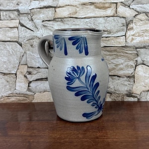 May include: A gray stoneware pitcher featuring a curved handle and a wide spout. The exterior is decorated with hand-painted cobalt blue floral patterns, including a large central flower motif and a series of smaller leaf designs around the rim.