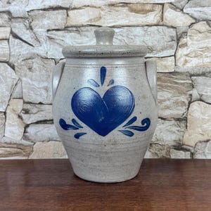 May include: A gray ceramic jar with a lid and two handles. The jar features a large blue heart design in the center, with blue accents around it. The jar is sitting on a wooden surface.