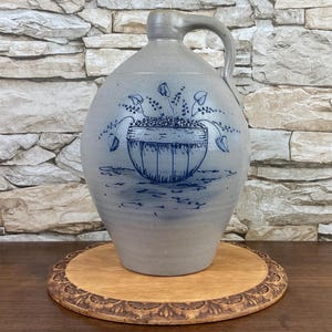 May include: A blue and white stoneware pitcher with a floral design. The pitcher has a wide base and a narrow neck. It has a handle on the side. The pitcher is sitting on a wooden base.
