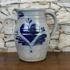 May include: A stoneware pitcher with a blue heart design. The pitcher has a light gray base color with a dark blue heart and floral accents. It features a curved handle and a spout for pouring. The pitcher is likely handmade and has a rustic, vintage aesthetic.