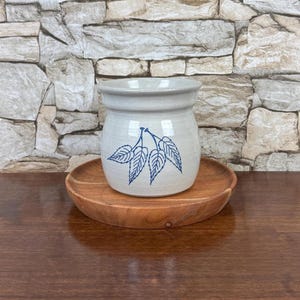 May include: A light gray ceramic crock with a wide rim and a blue line drawing of three leaves on the front, resting on a circular wooden plate. The crock has a smooth, glossy finish and a rounded body shape, sitting centered on the wood base.