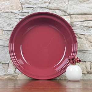 May include: A large, round, burgundy-colored ceramic serving bowl. The bowl has a glossy finish and is set on a wooden surface. A small white vase with burgundy flowers is next to the bowl.