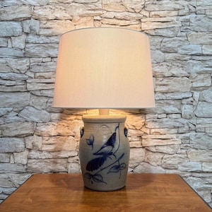 May include: A stoneware lamp with a beige lampshade. The lamp base is a gray-blue color with a blue bird and leaf design. The lamp sits on a wooden table against a stone wall background.