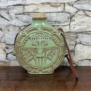 May include: A green ceramic flask with a cork stopper and a brown leather strap. The flask features a detailed embossed design of a bird. The flask is approximately 6 inches tall.
