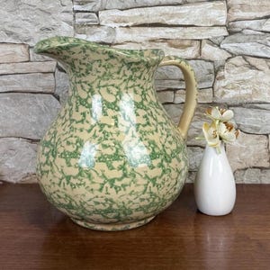 May include: A large, cream-colored ceramic pitcher with a green splatter pattern. The pitcher has a curved handle and a ruffled rim. A small white vase with faux flowers sits beside the pitcher.