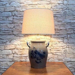 May include: A table lamp featuring a beige fabric drum shade and a ceramic base shaped like a two-handled crock. The light-colored ceramic base is decorated with a dark blue hand-painted illustration of a bird perched on a flowering branch.