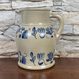 May include: A beige ceramic pitcher with a handle and a wide spout. The pitcher is decorated with blue floral patterns around the body. The pitcher sits on a wooden surface against a stone wall background.