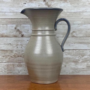 May include: A handmade stoneware pitcher with a light gray body and a dark blue rim and handle. The pitcher has a spout for easy pouring and a curved handle for comfortable grip. The ceramic pitcher is a decorative and functional item.