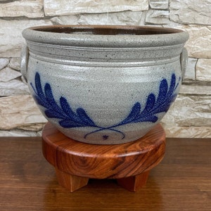 May include: A gray stoneware pot with blue leaf designs and two handles. The pot is sitting on a wooden stand with three legs.