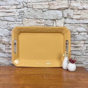 May include: A rectangular mustard yellow serving tray with integrated side handles and a raised border featuring a subtle grid pattern. Two small white ceramic vases are positioned in the lower right corner, one containing dried flowers and the other a small red bloom.