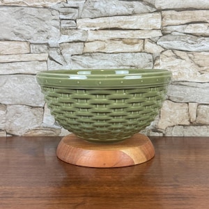 May include: A sage green ceramic bowl with a textured exterior that mimics the appearance of a woven basket. The bowl features a smooth, flat rim with small dot detailing and is supported by a circular, light-toned wooden base stand.