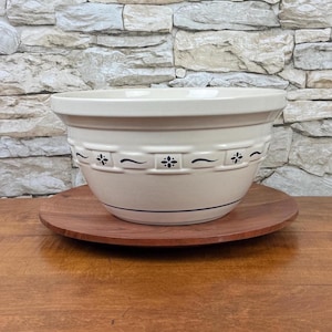 May include: Cream-colored ceramic mixing bowl with a decorative blue floral and scroll design. The bowl sits on a round, wooden serving tray. The background is a stone wall.