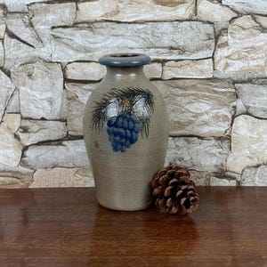 May include: A beige ceramic vase with a narrow neck and a blue rim, featuring a hand-painted blue pinecone and pine needle design on the front. A single brown pinecone is positioned next to the base of the vase on a wooden surface.