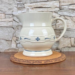 May include: A cream-colored ceramic pitcher featuring a rounded body, a wide pouring spout, and a curved handle. The lower section is decorated with a repeating blue pattern of small floral motifs and horizontal lines.