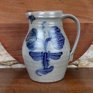 May include: A gray stoneware pitcher with a blue and white floral design. The pitcher has a wide mouth and a handle.