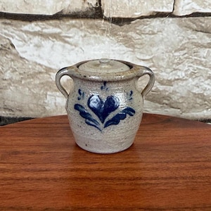 May include: A small, speckled beige stoneware crock with a matching lid and two side handles. The front of the vessel features a hand-painted cobalt blue heart design with decorative leaf-like accents extending from the base of the heart shape.
