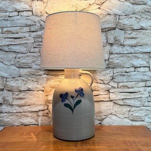 May include: A stoneware lamp with a beige linen lampshade. The lamp base is a gray jug with a handle and blue floral design. The lamp sits on a wooden surface against a stone wall background.