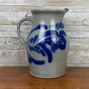 May include: A gray ceramic pitcher with a blue floral design. The pitcher has a handle and a spout. The blue floral design is painted on the front of the pitcher. The pitcher is sitting on a wooden surface.