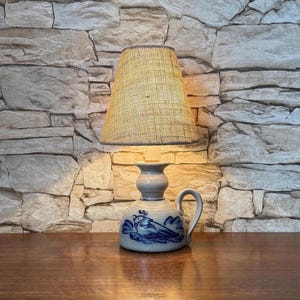 May include: A table lamp with a beige woven fabric lampshade and a light gray ceramic base. The base features a blue hand-painted bird illustration, a rounded body, a narrow neck, and a curved handle on the side, emitting a warm yellow light.