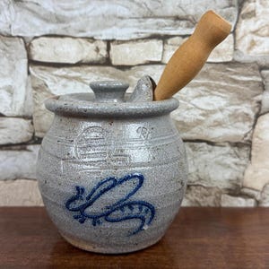 May include: A gray ceramic honey pot with a lid and a wooden honey dipper. The pot has a blue bee design and the words "Pottery Works" are visible. The pot is sitting on a wooden surface.
