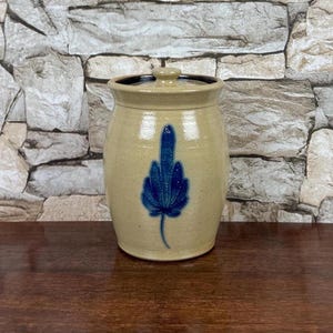 May include: A beige ceramic stoneware crock with a cylindrical body and a matching lid. The exterior features a speckled finish and a prominent, hand-painted cobalt blue leaf design centered on the front of the vessel.