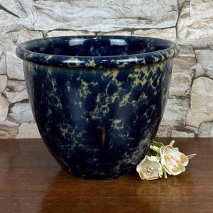 May include: A dark blue ceramic planter with a speckled yellow pattern. The planter has a rounded shape and a slightly flared rim. The planter is sitting on a wooden surface.