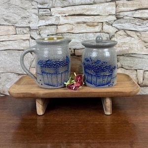 May include: A gray ceramic pitcher and a matching lidded jar, both featuring a blue embossed design of a basket filled with berries. The two pieces are displayed on a small wooden riser, with a small artificial flower placed between them.