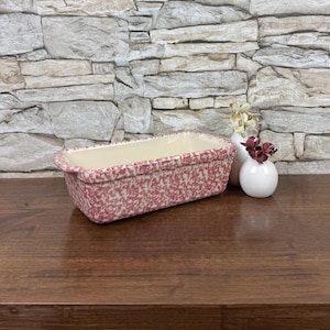 May include: A rectangular ceramic loaf pan with a speckled red and white pattern. The interior is a cream color. Two small white vases with flowers are next to the pan. The pan is on a wooden surface.