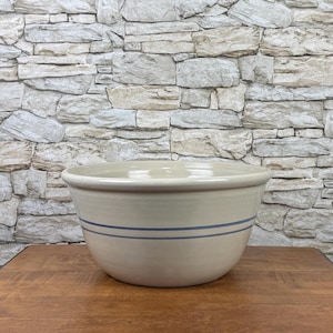 May include: A large, round, cream-colored ceramic mixing bowl featuring two thin, horizontal blue stripes encircling the exterior. The bowl has a smooth, glossy finish with a wide, slightly flared rim and a deep, curved basin for food preparation.