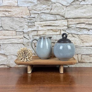 May include: A light blue ceramic creamer and a matching lidded sugar bowl with a dark brown lid, positioned on a small rectangular wooden riser alongside a decorative dried flower bloom, showcasing a speckled glaze finish.