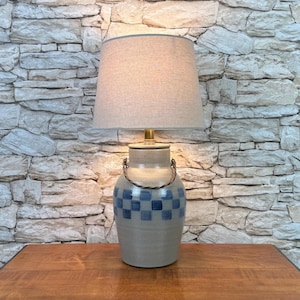 May include: A table lamp featuring a beige ceramic base with a blue checkered pattern and a twisted metal handle. The lamp is topped with a tapered, light-colored fabric lampshade that emits a soft glow, resting on a brass-colored metal neck.