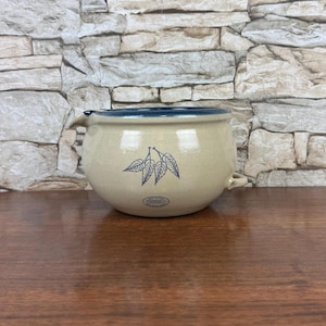 May include: A beige stoneware bowl with a dark blue rim and handles. The bowl features a blue leaf design and a small, round label. The background is a stone wall.